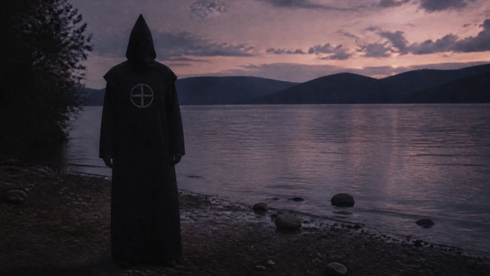 Hooded figure wearing a black executioner-style hood with a circle-and-cross bib standing near a lakeshore at dusk